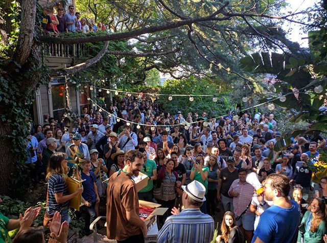 A view framed by trees of a party where a few hundred people look towards the camera and stage where prizes are given out. The image has light strung between the trees framing the shot.